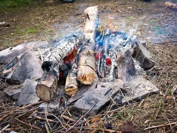 Kleine Flamme zwischen Holzscheiten und Steinen, Rauch steigt auf
