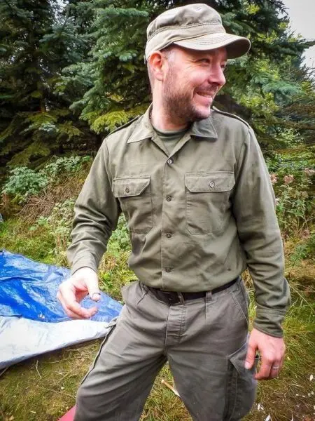 Mann in militärischer Kleidung steht neben einer blauen Plane im Wald