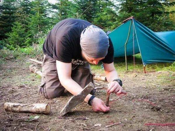 Mann kniet und bearbeitet Holzstück mit einem Steinwerkzeug