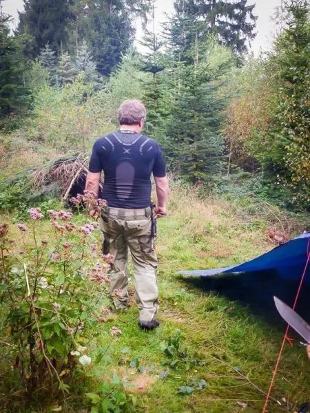 Mann mit Ausrüstung geht durch bewaldetes Gelände in Richtung eines blauen Tarps