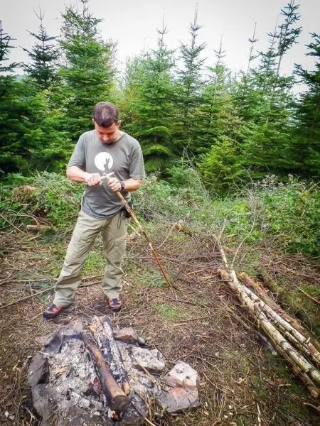 Mann schnitzt mit einem Messer an einem Stock neben einem kleinen Feuer