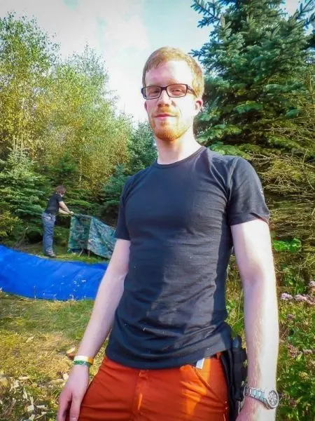 Mann steht in der Natur, im Hintergrund wird eine Tarp-Notunterkunft aufgebaut