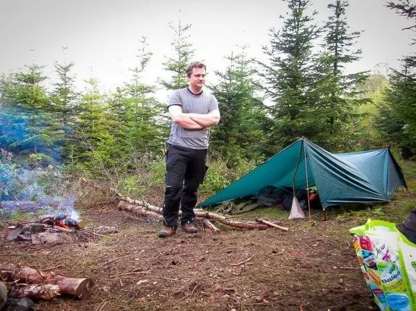Mann steht neben einer Tarp-Notunterkunft und einem kleinen Feuer im Wald