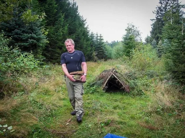 Mann trägt Holzstück zur selbstgebauten Tarp-Notunterkunft im Wald
