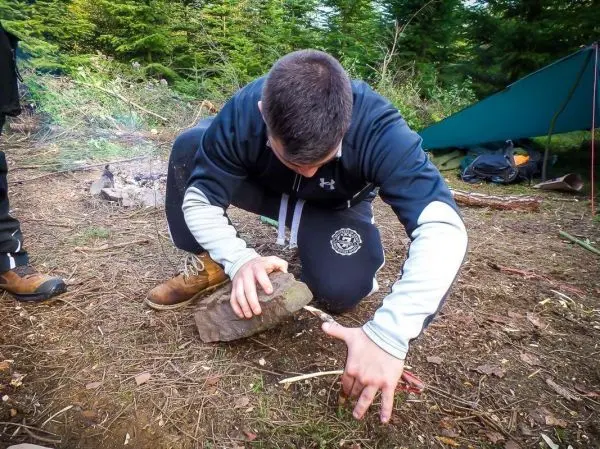 Person bearbeitet Holzstück mit einem Stein beim Survival Training