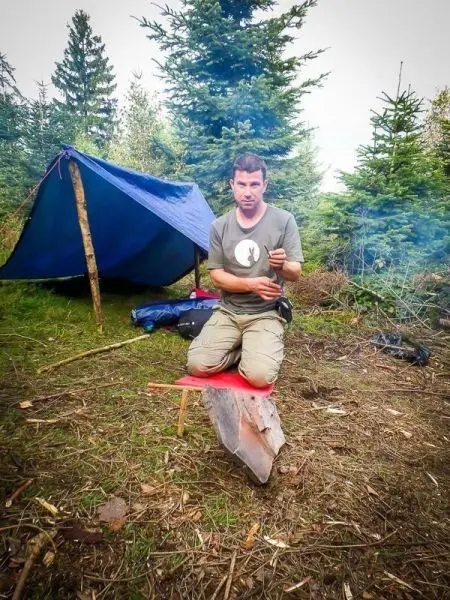 Person sitzt auf einem Holzscheit und hält ein Messer in der Hand
