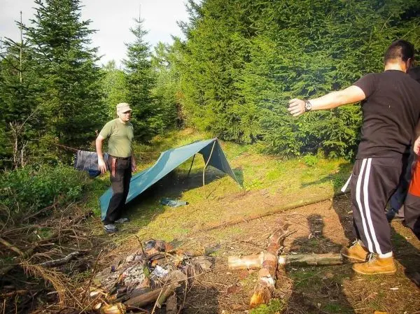 Person steht neben Zelt und beobachtet Feuerstelle beim Überlebenstraining