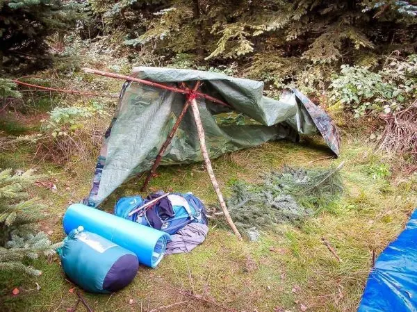 Tarp-Notunterkunft aus einer Plane und Ästen, daneben Rucksäcke und Isomatten