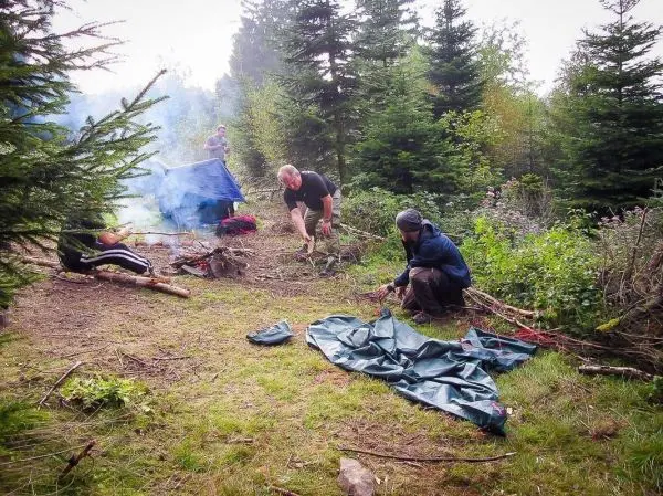 Zwei Personen sammeln Äste, während eine Tarp-Notunterkunft im Hintergrund sichtbar ist