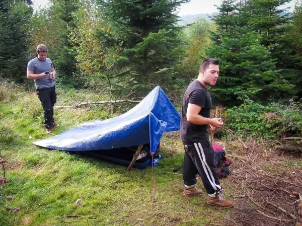 Zwei Personen stehen neben einer Tarp-Notunterkunft im Wald