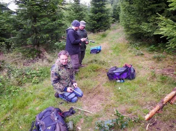 Drei Personen mit Rucksäcken stehen und knien auf einem Waldweg