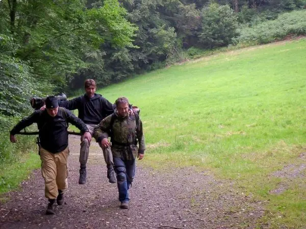 Drei Personen tragen eine improvisierte Trage mit einem verletzten Mann über einen Weg