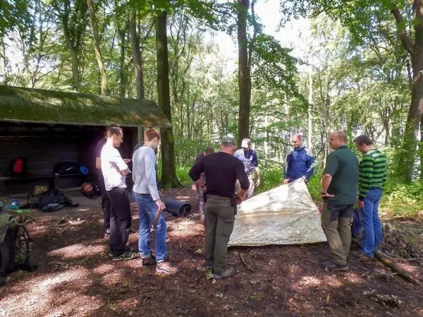 Gruppe baut Tarp-Notunterkunft im Wald auf, umgeben von Bäumen