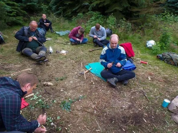 Gruppenteilnehmer bearbeiten Holzstücke mit Messern auf dem Boden in einem Waldgebiet