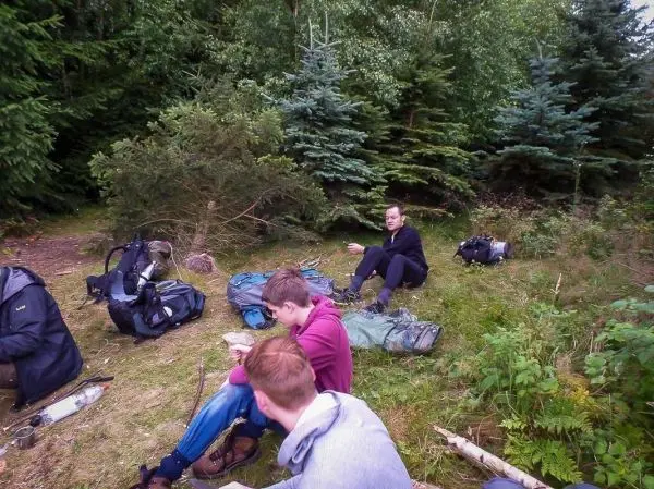 Gruppierung von Rucksäcken und Ausrüstung im Wald, Teilnehmer sitzen am Boden