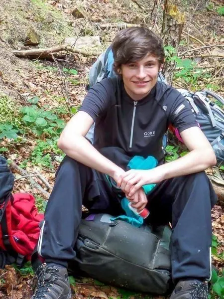 Junge sitzt auf einem Rucksack im Wald, umgeben von Bäumen und Laub