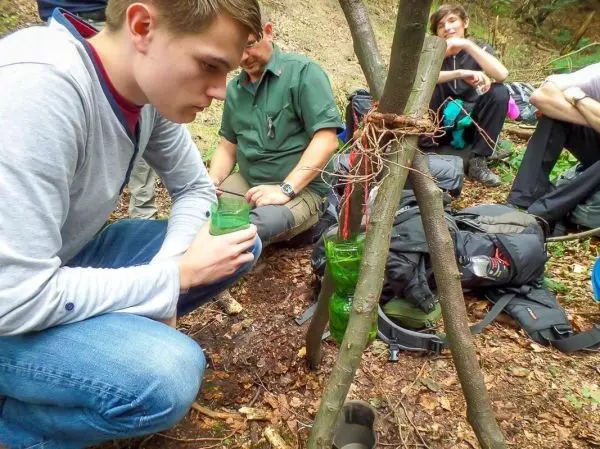 Junger Mann beobachtet selbstgebaute Wasserfilterkonstruktion aus Ästen und Flaschen