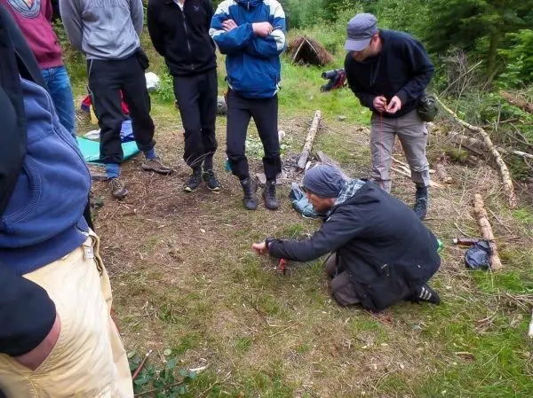 Mann zündet Zunder mit Feuerstahl auf dem Boden an, umringt von Zuschauern