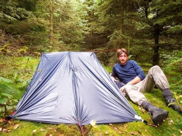 Tarp-Notunterkunft steht im Wald, Person sitzt daneben auf dem Boden