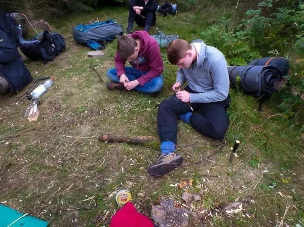 Zwei Personen schnitzen mit Messern an Holzstöcken auf dem Boden