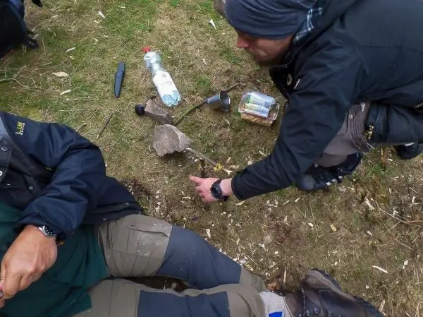 Zwei Personen sitzen auf dem Boden, einer zeigt auf einen Stein und eine Flasche