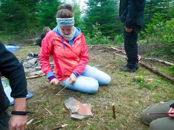 Frau schnitzt mit einem Messer an einem Holzstück auf dem Boden