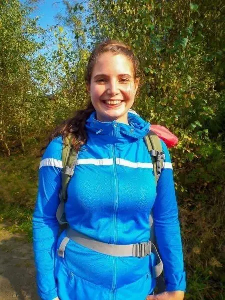 Lächelnde Person in blauer Jacke mit Rucksack vor grüner Vegetation