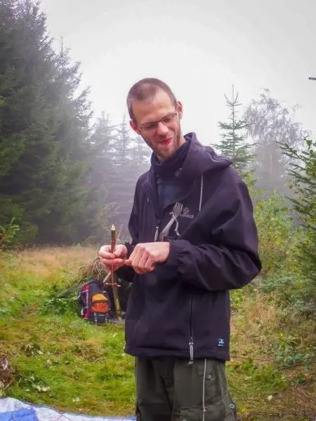 Mann hält einen Pfeil in der Hand und schaut darauf, umgeben von Bäumen