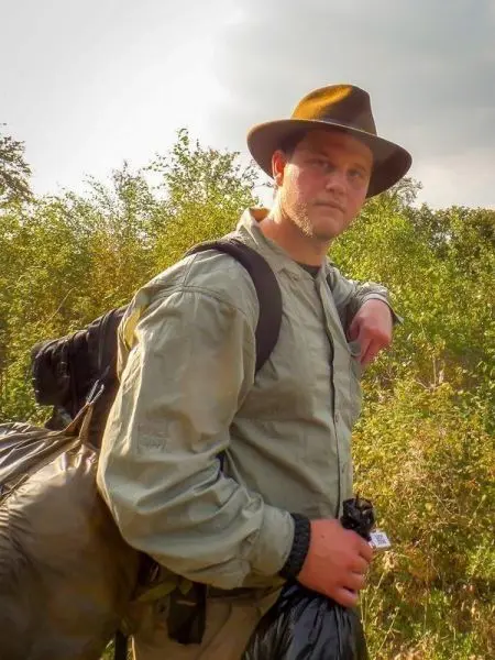 Mann mit Hut und Rucksack steht vor dichter Vegetation