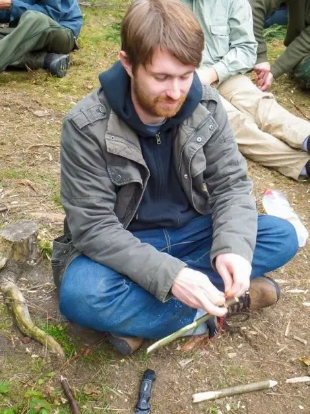 Mann sitzt im Gras und schnitzt mit einem Messer an einem Holzstück