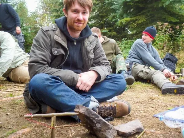 Mann sitzt im Schneidersitz neben einer Holzhebel-Konstruktion mit Stein