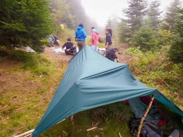 Tarp-Notunterkunft ist auf dem Boden gespannt, mehrere Personen arbeiten im Hintergrund