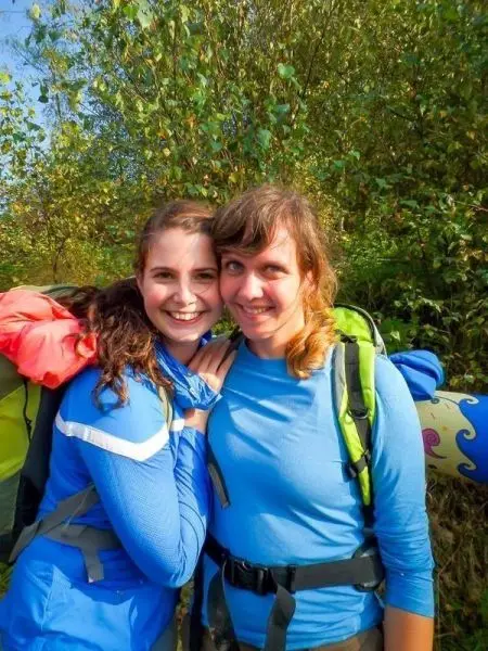 Zwei Frauen mit Rucksäcken stehen lächelnd vor einer grünen Kulisse