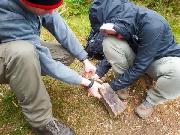 Zwei Personen bearbeiten einen Holzblock mit einem Messer auf dem Boden