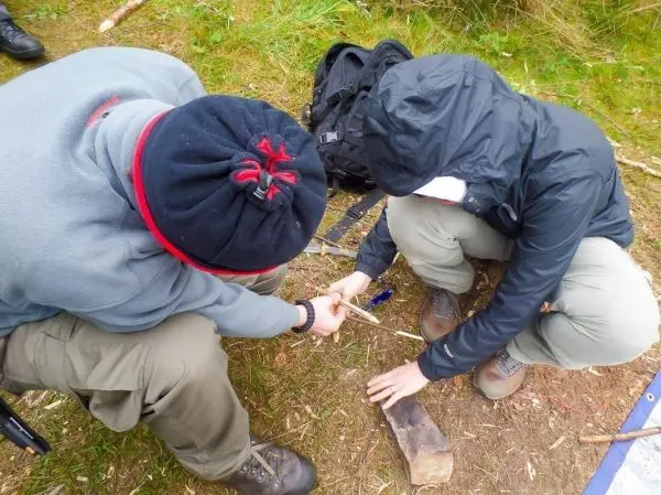 Zwei Personen bearbeiten einen Holzstock mit einem Stein auf dem Boden