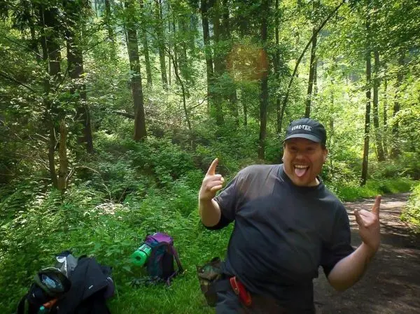 Ein Mann posiert fröhlich im Wald, Rucksack und Ausrüstung liegen am Boden