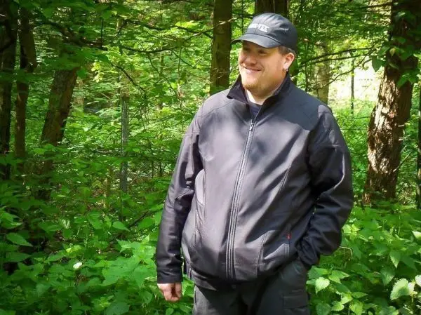 Ein Mann steht lächelnd im Wald zwischen Bäumen und grünen Pflanzen