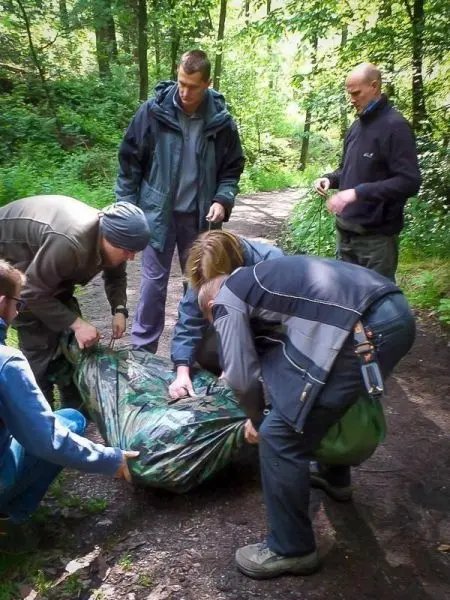Fünf Personen heben eine camouflagefarbene Tarp-Notunterkunft auf einem Waldweg