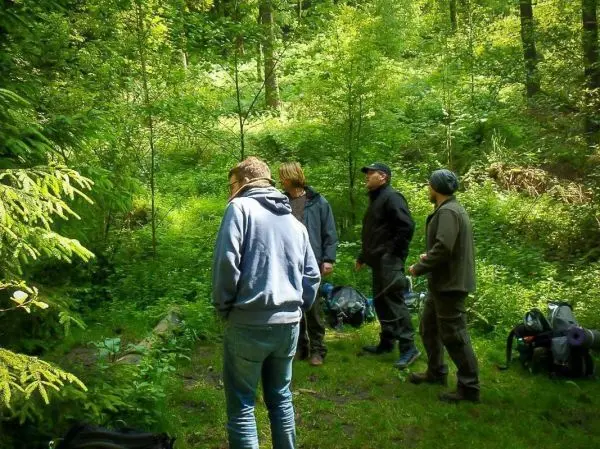 Fünf Personen stehen im Wald und betrachten die Umgebung, Rucksäcke liegen am Boden