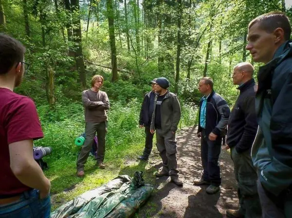 Gruppe diskutiert im Wald über Überlebenstechniken, Tarp liegt auf dem Boden