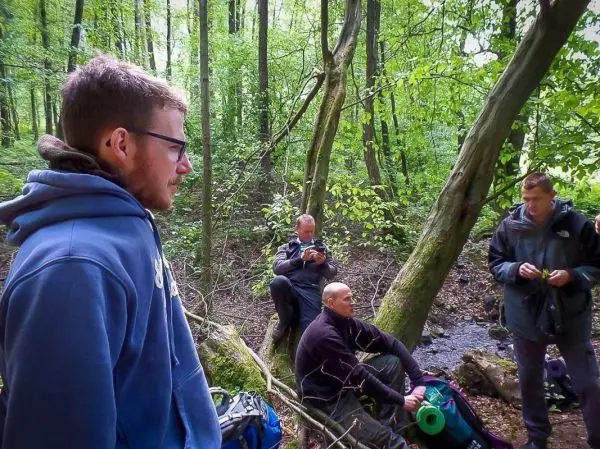 Gruppe sitzt im Wald, Rucksäcke liegen auf dem Boden, Wasserlauf im Hintergrund