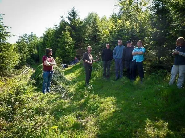 Gruppe steht auf einem Waldweg, eine Person erklärt mit Werkzeug in der Hand