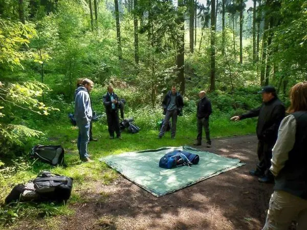 Gruppe steht um eine grüne Tarp-Notunterkunft mit Rucksäcken im Wald
