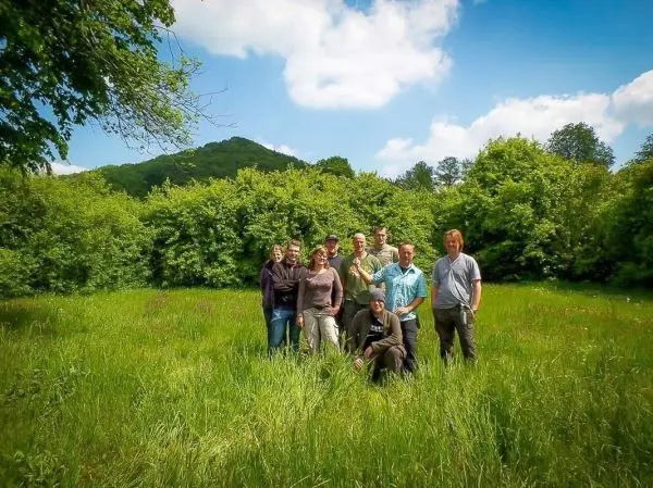 Gruppe von acht Personen steht in einer Wiese umgeben von Bäumen