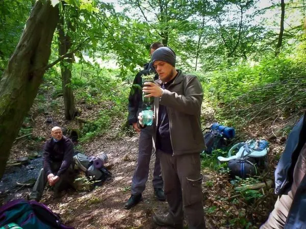 Mann hält ein Glas mit Wasser in einem Waldgebiet, Rucksäcke liegen am Boden