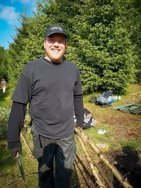 Mann hält ein Messer und steht neben einem Holzstapel im Wald