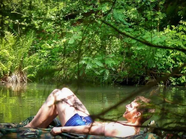 Mann liegt entspannt auf einer Matte am Ufer eines Gewässers, umgeben von Pflanzen