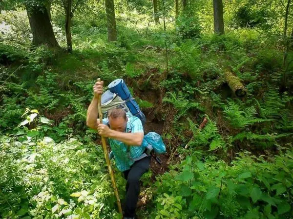 Mann mit Rucksack erklimmt steilen Hang mit einem Stock zur Unterstützung