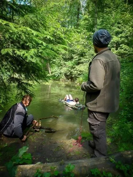 Mann mit Stock am Ufer, ein weiterer Mann auf improvisierter Schwimmkonstruktion im Wasser