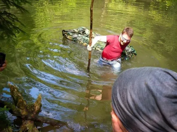 Mann mit Stock überquert seichtes Wasser, während er eine camouflagefarbene Decke zieht